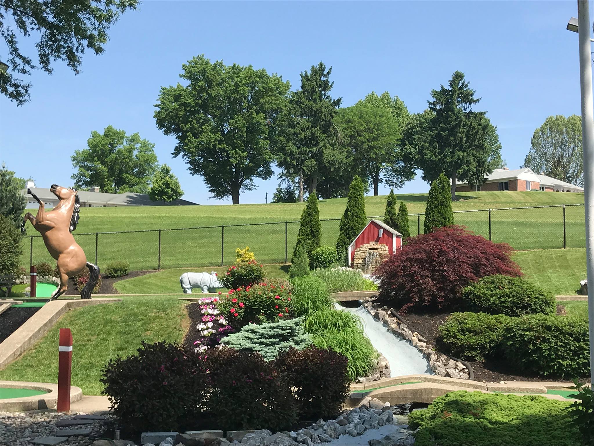 A mini-golf course featuring a rearing horse statue, colorful flowers, a small water stream, manicured bushes, a red barn replica, and green trees under a clear blue sky.