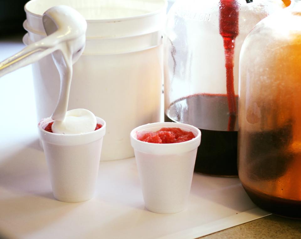 Two white foam cups are being filled with red shaved ice; one cup is topped with a swirl of white cream from a ladle. Large plastic jugs with red syrup are in the background.