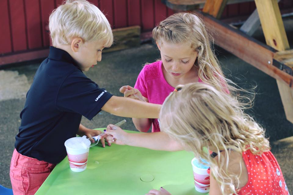 Three young children sit at a green table, playing and reaching for something. Each has a cup in front of them. The boy wears a black shirt and the two girls have wavy blonde hair and wear pink and red dresses.