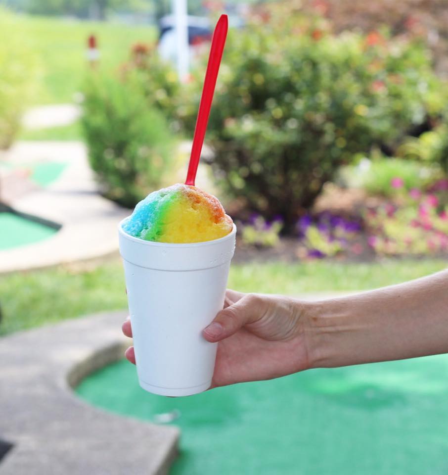 A hand holds a white styrofoam cup filled with rainbow-colored shaved ice and a red spoon, with a green and landscaped outdoor area in the background.
