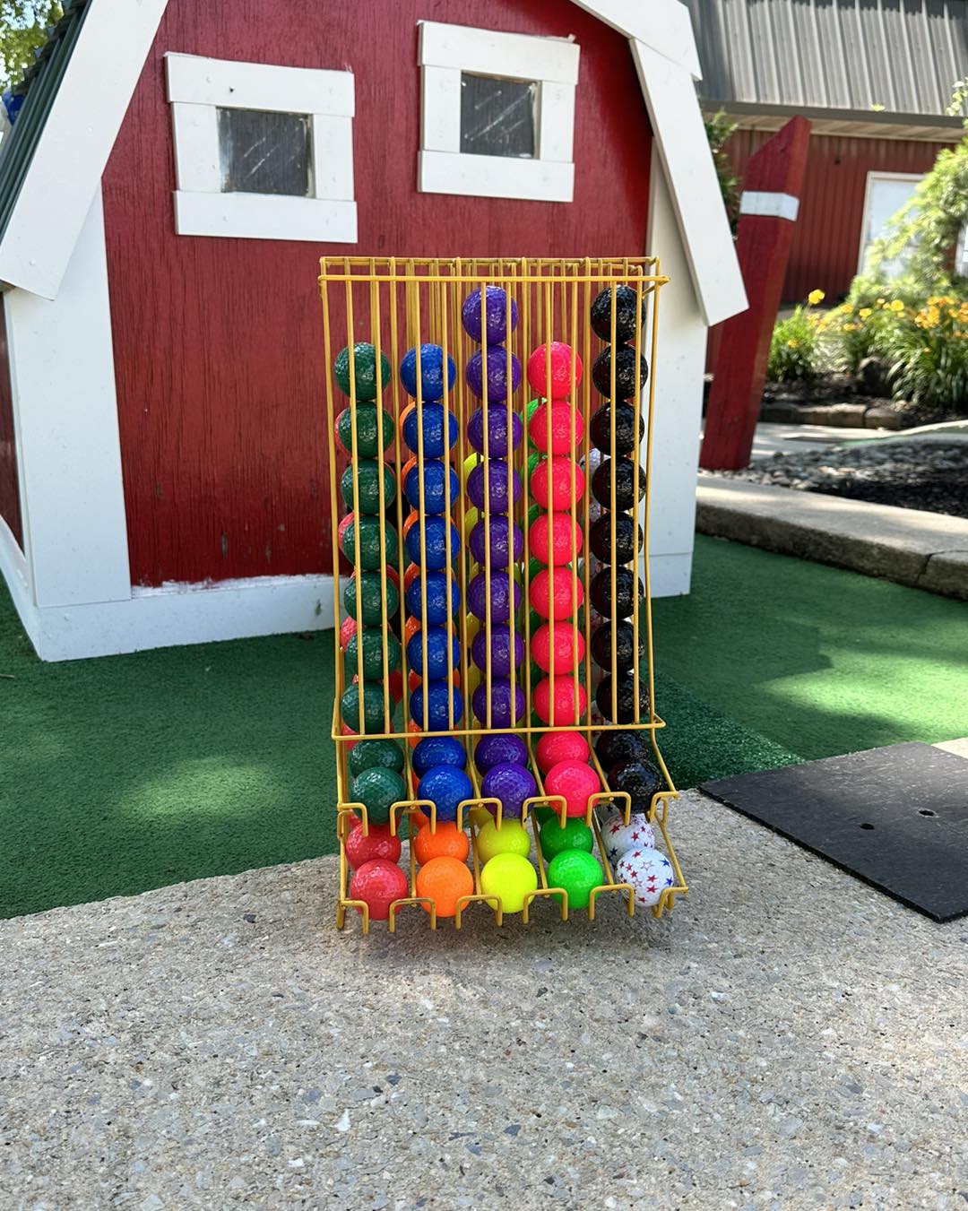 A metal rack displays colorful mini-golf balls arranged in rows in front of a red and white barn structure on a mini-golf course. The balls are grouped by color, including blue, green, orange, pink, yellow, and black.