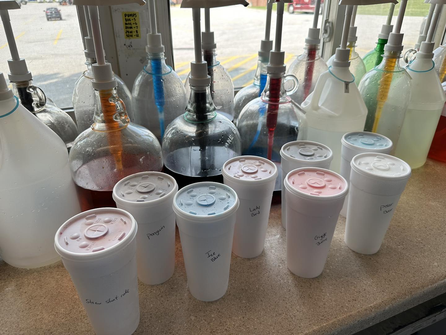 Several large syrup dispensers with pumps are lined up on a counter behind five white styrofoam cups with handwritten labels, each with a plastic lid. The scene appears to be at a snow cone or drink stand.