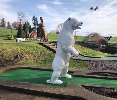 A large white polar bear statue stands on a mini-golf course, with a moose statue and smaller animal figures in the background surrounded by green grass and trees.