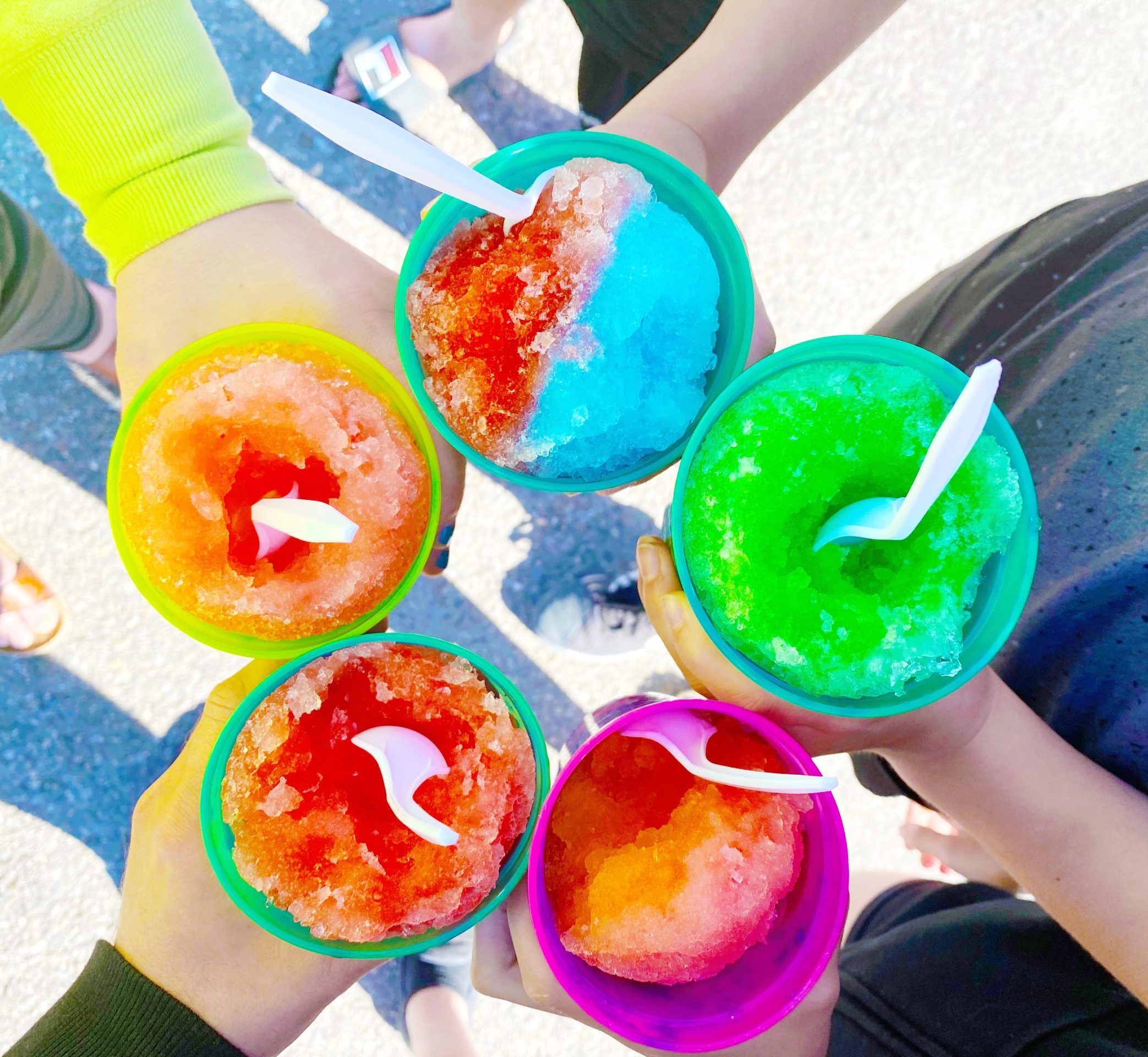 A top-down view of five colorful shaved ice cups with plastic spoons, held by different people standing outdoors on a sunny day.