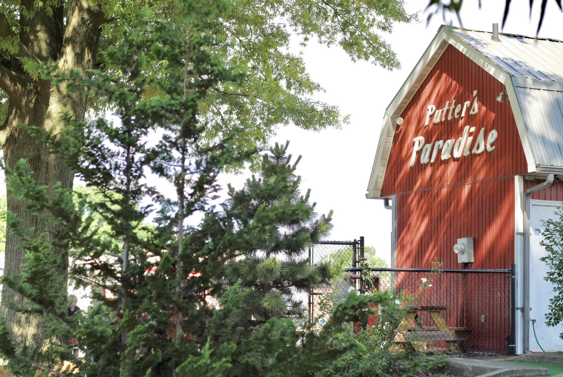 A red barn-like building with the words "Putter's Paradise" painted on it is partially hidden behind a tree and greenery. There is a picnic table and a metal fence near the building.