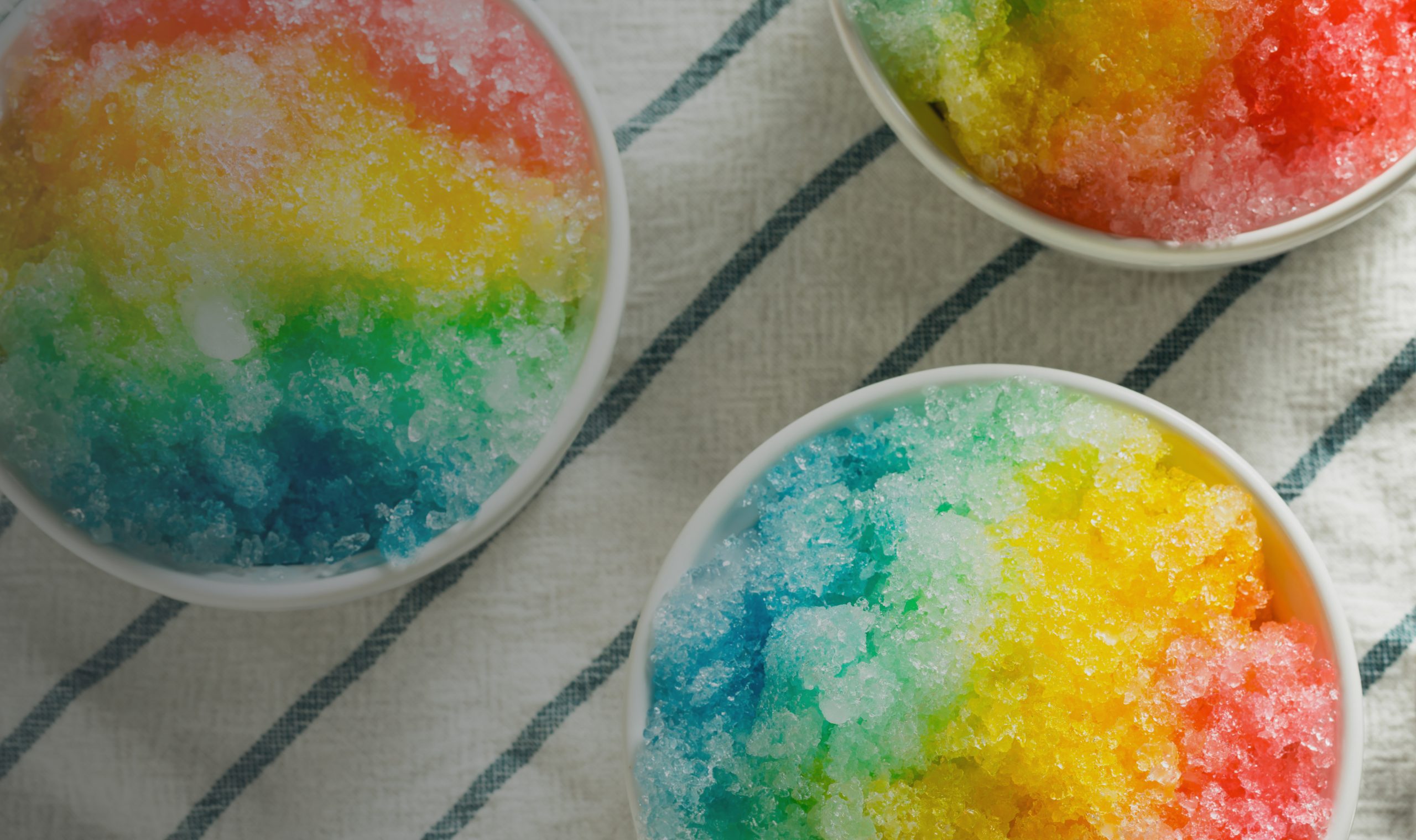 Three bowls of shaved ice with vibrant rainbow-colored syrup sit on a white towel with dark blue stripes. The ice displays shades of red, yellow, green, and blue, creating a bright and colorful scene.
