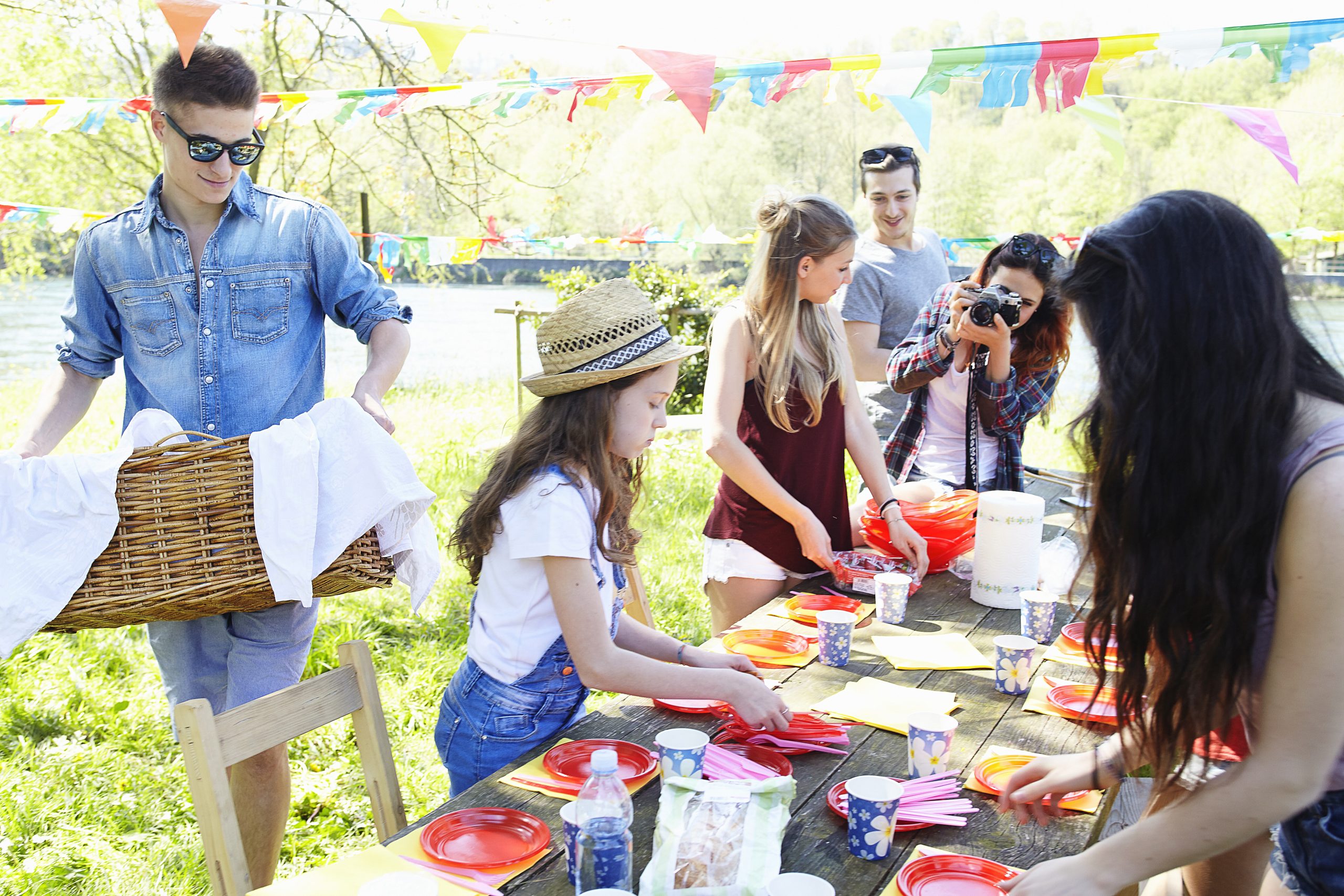 A group of young people set up a picnic table outdoors, arranging plates and napkins. Bright bunting hangs above, and one person takes a photo while another carries a wicker basket. The scene is cheerful and sunny.