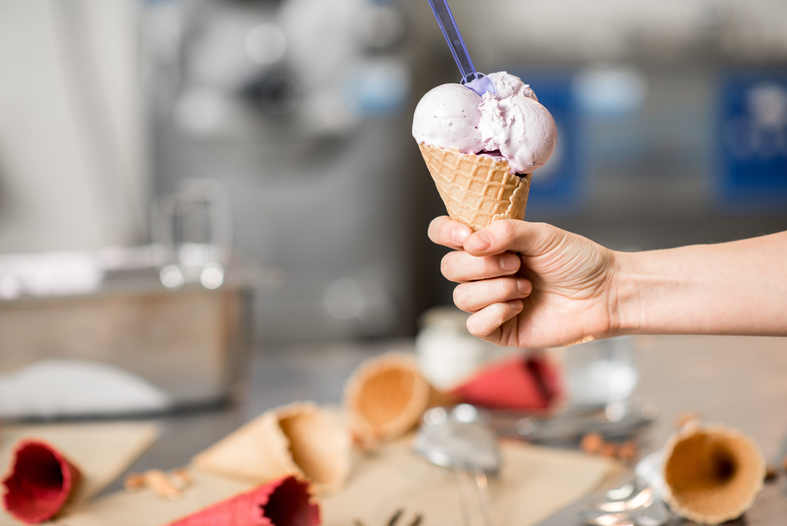 A hand holding an ice cream cone with two scoops of lavender-colored ice cream, with empty cones and utensils scattered on a table in the blurred background, reminds one of reading Terms and Conditions before indulging in any Website Policy.