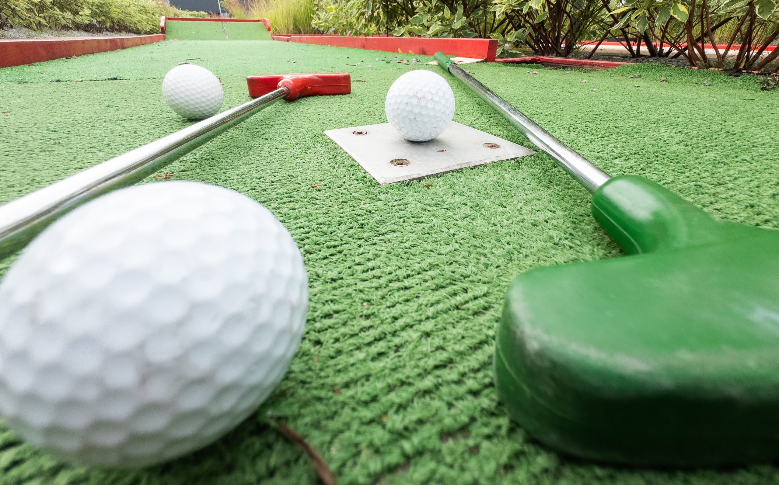 Close-up view of a mini-golf course with two putters, one red and one green, next to three white golf balls on artificial green turf, surrounded by low red borders and greenery—please review our Privacy Policy for more information.