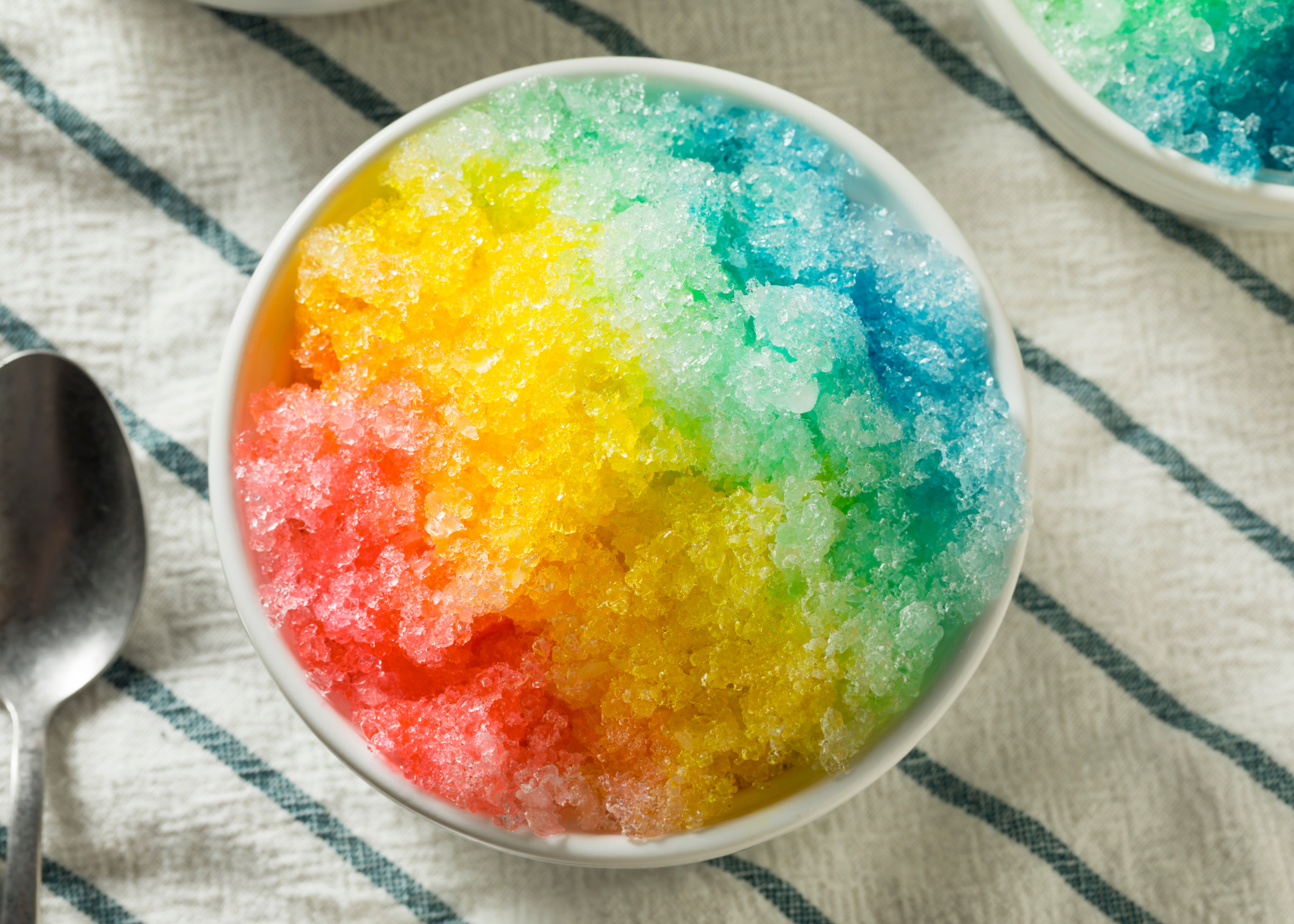 A white bowl filled with rainbow-colored shaved ice, showing red, yellow, green, and blue sections, sits on a white cloth with blue stripes. A metal spoon is placed nearby.