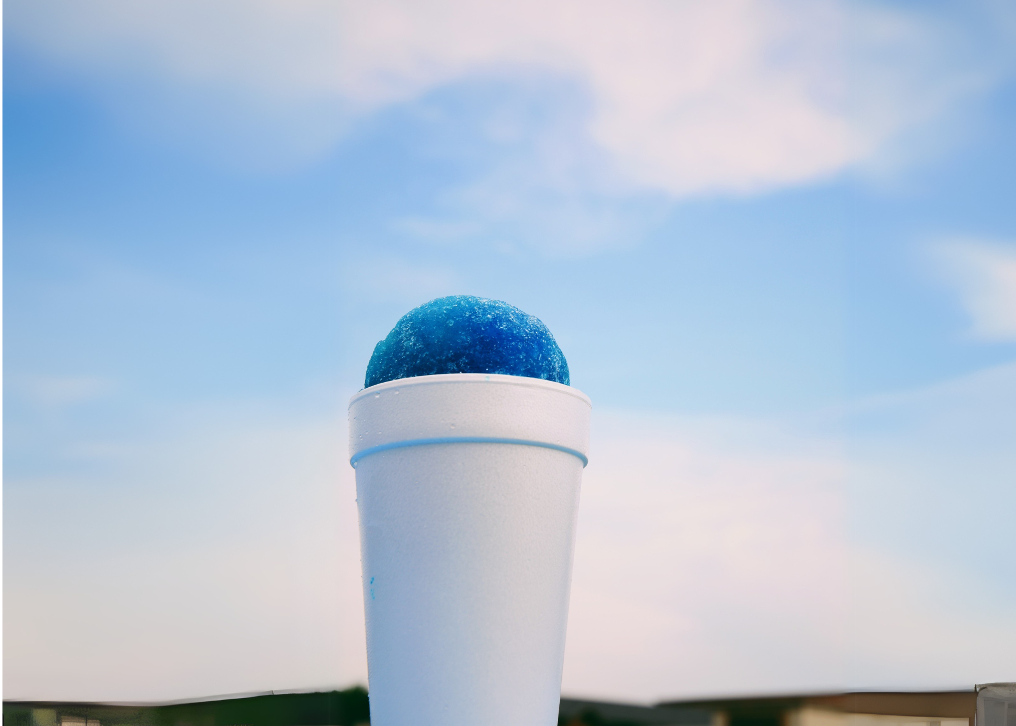 A white styrofoam cup filled with blue shaved ice is held up against a bright, partly cloudy sky with blurred buildings in the background.