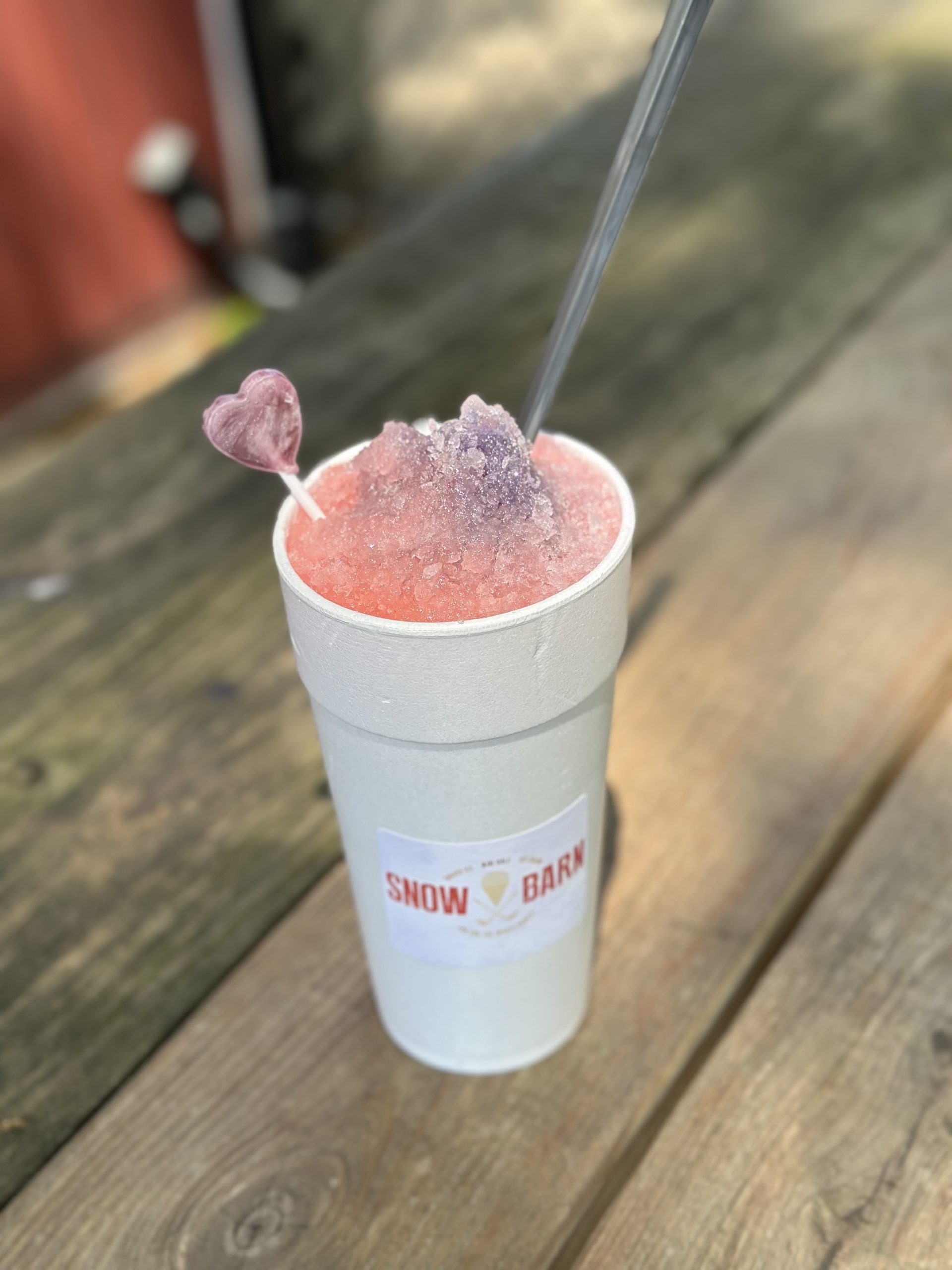 A white cup labeled “Snow Barn” holds a pink and purple snow cone with ice crystals, a clear straw, and a plastic heart-shaped stir stick, sitting on a wooden picnic table outdoors.